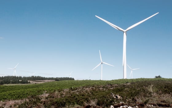 Image of a wind turbine located on a hill, in a green landscape with blue skies above.